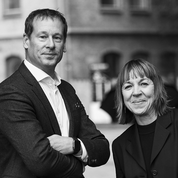 Svartvit porträttbild av Jonas Westerén och Charlotte Sigeskog i affärskläder, stående utomhus framför en suddig bakgrund med tegelbyggnader. Mannen har armarna i kors och bär en kavaj med en vit skjorta, medan kvinnan ler och bär en svart kavaj över en mörk tröja.
