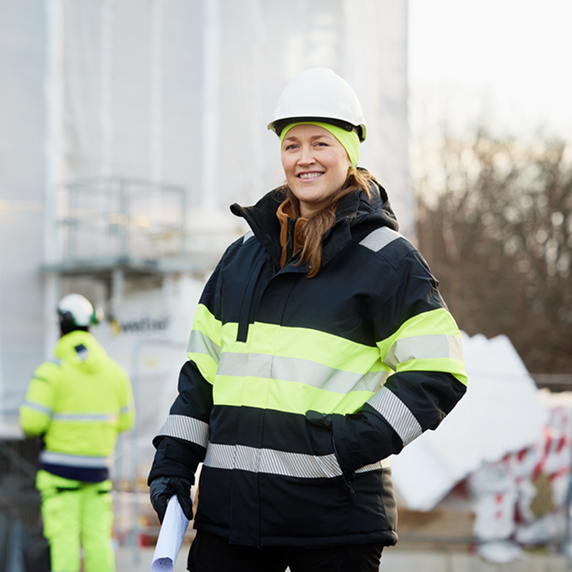 Leende kvinna i skyddsutrustning, inklusive reflexjacka och hjälm, står på en byggarbetsplats med ritningar i handen. I bakgrunden syns en annan arbetare och byggmaterial.