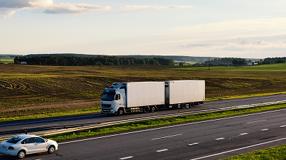 Lastbil med dubbla släp kör på en motorväg genom ett öppet landskap, med en personbil i motsatt körfält och fält i bakgrunden.